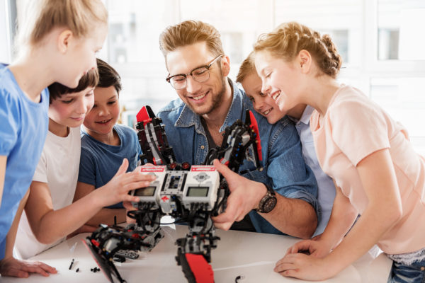 Cheerful adult man is surrounded by interested young technicians. He showing robot to children. They are sitting near desk